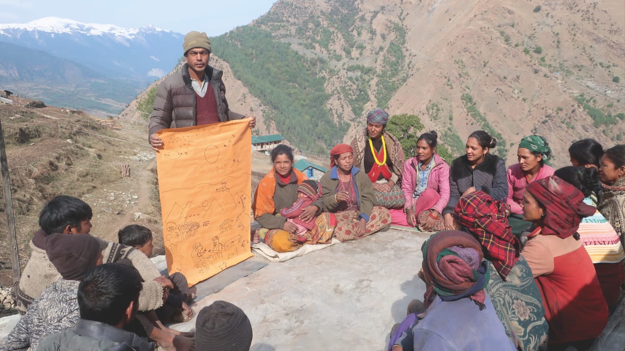 Group of women and children on a mountain side listen to presentation given by a man holding a drawing on orange paper.
