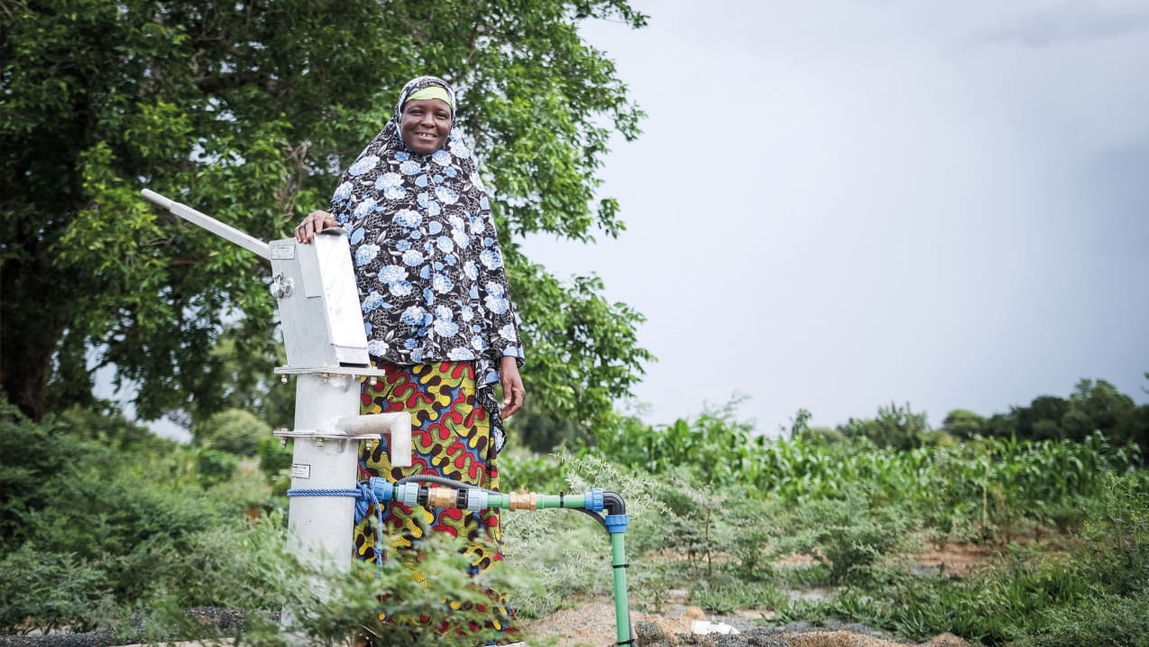 A woman smiles and stands with her hand on top of a water pump. Behind her is a tree and green shrubs.