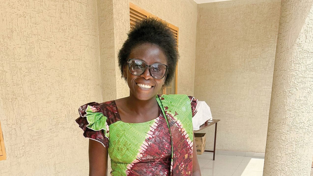A smiling woman with glasses and traditional dress stands in front of a textured wall.