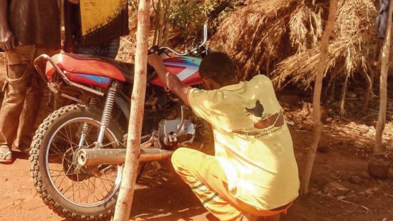 Person repairs a motorcycle in a rustic, rural setting.