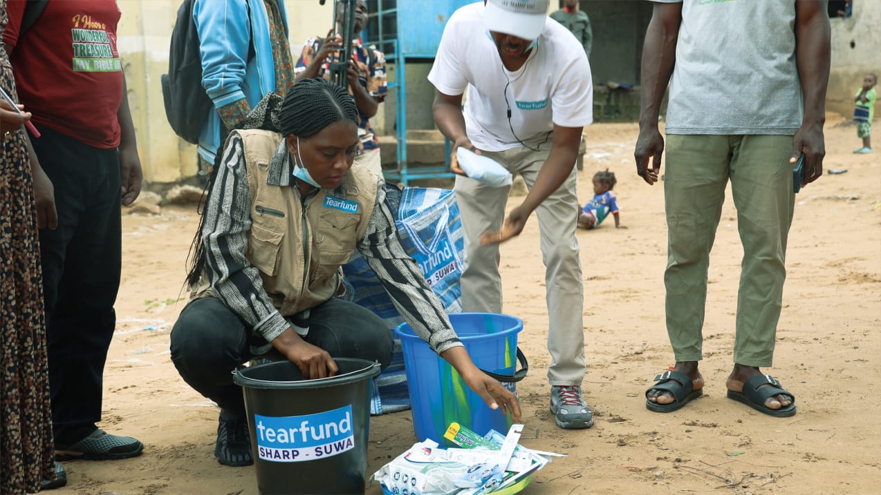 Two people dressed in clothing with the Tearfund logo crouch to sort items into branded buckets outdoors with others nearby.