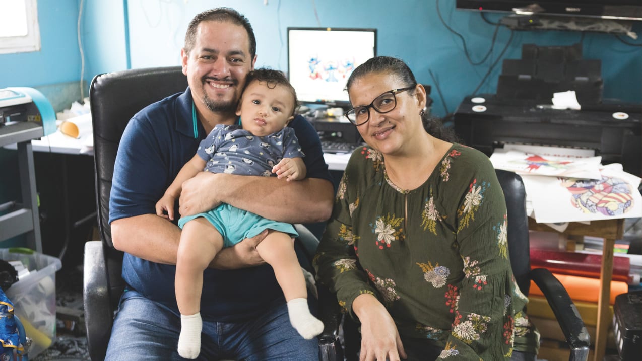 Smiling family of three (man, woman, baby) in a home office or workshop setting.