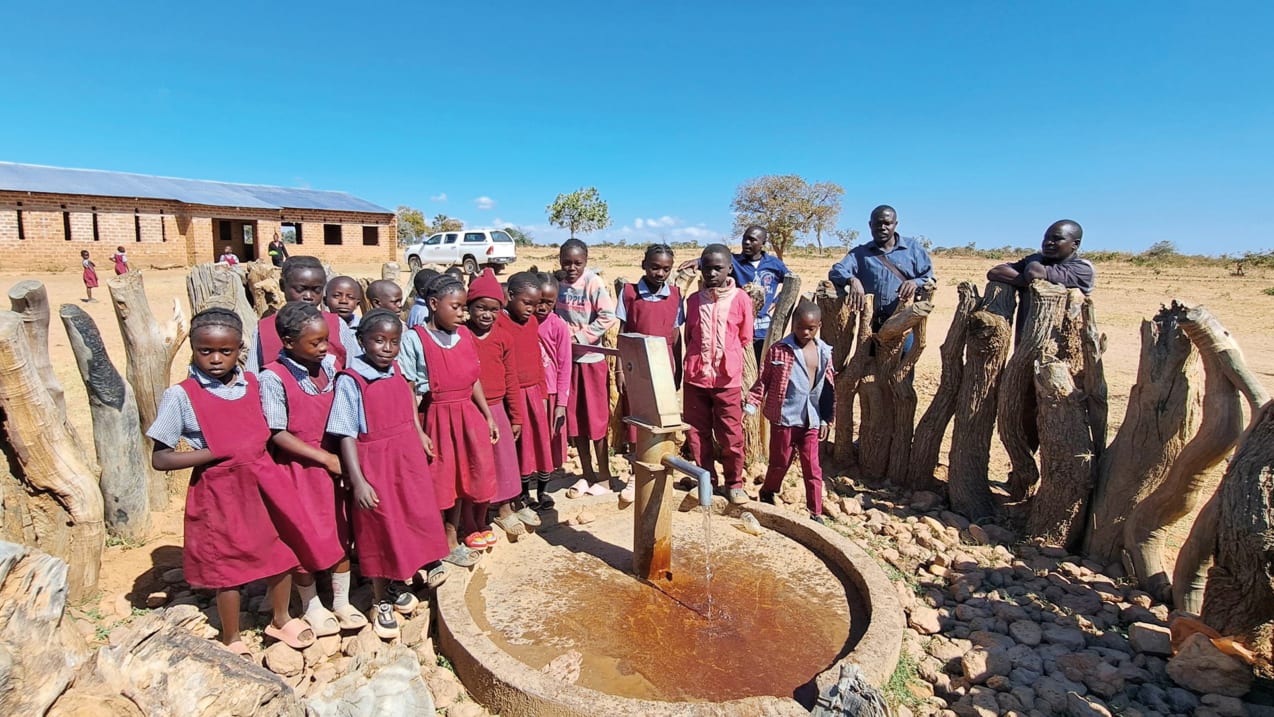 A group of school children in red uniforms stand around a hand water pump, with water flowing into a concrete basin. A brick school building is in the background under a clear blue sky.