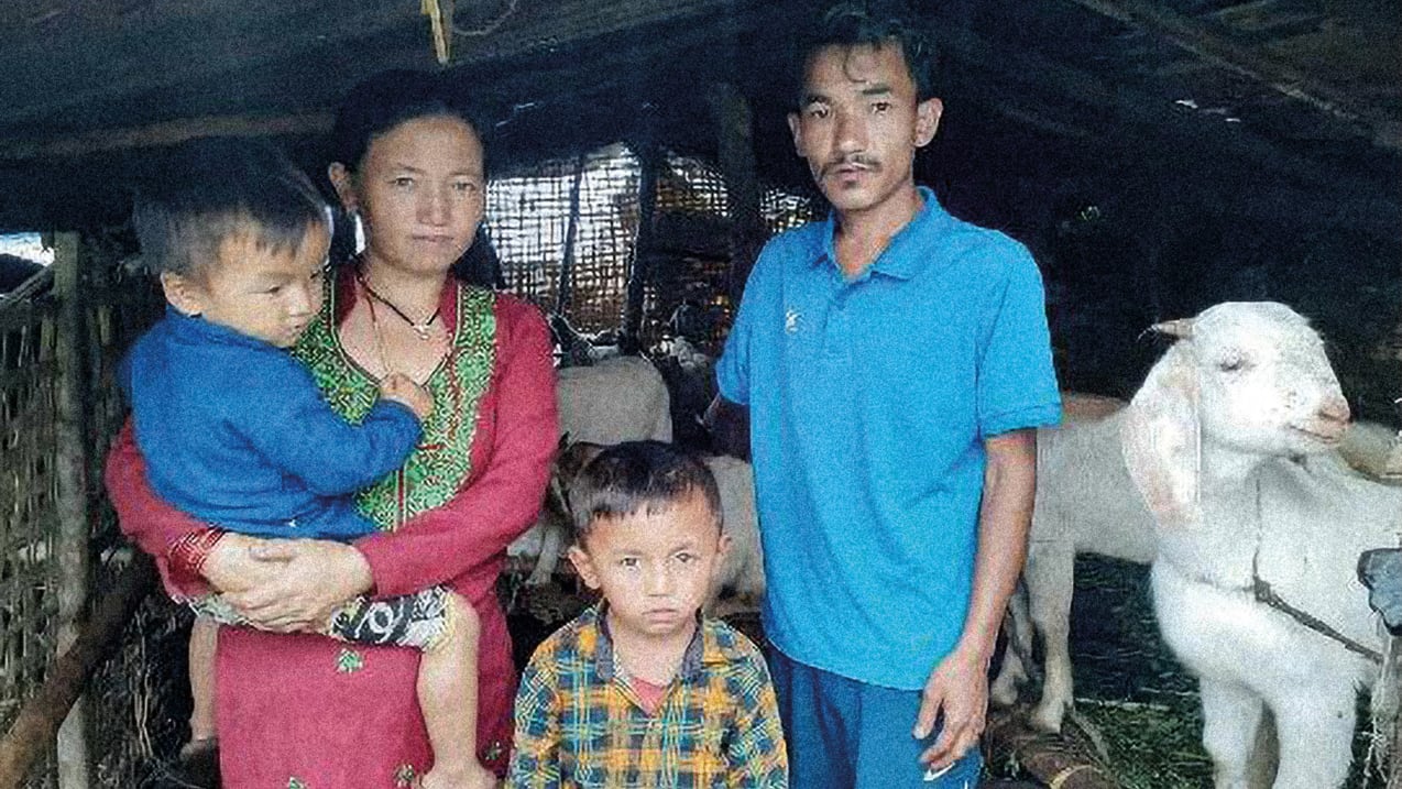 A family of four stands in a rustic animal shelter. A woman in a red garment holds a baby in a blue top, next to a man in a blue polo shirt. Another child in a plaid shirt stands in front, with several white goats visible behind them.