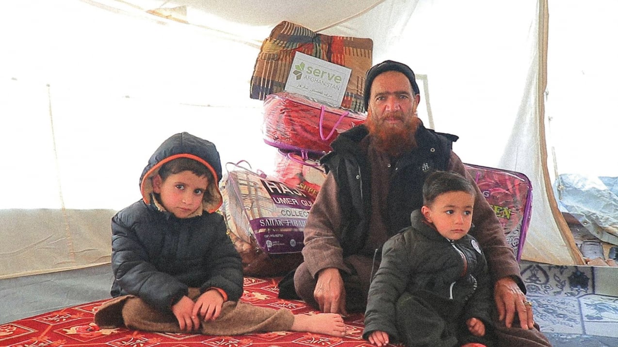 An adult man with a reddish beard and two young boys sit on a red patterned rug inside a white tent. Behind them are stacked aid packages, including blankets, with a "Serve Afghanistan" logo visible.