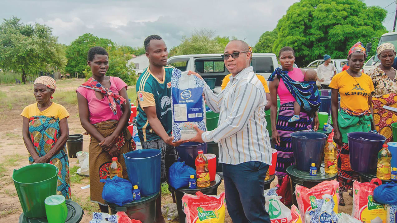A man in a striped shirt hands a large bag of rice to another man, surrounded by women and children, with buckets and food supplies arranged on the ground.