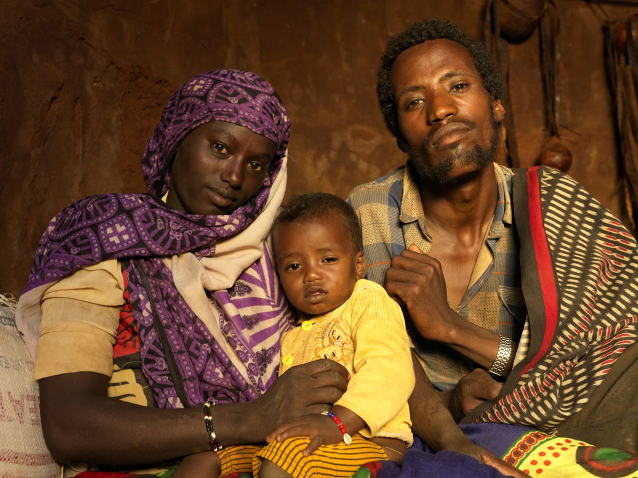 A woman wearing purple, holding a baby in a yellow cardigan, sits beside a man in a checked shirt
