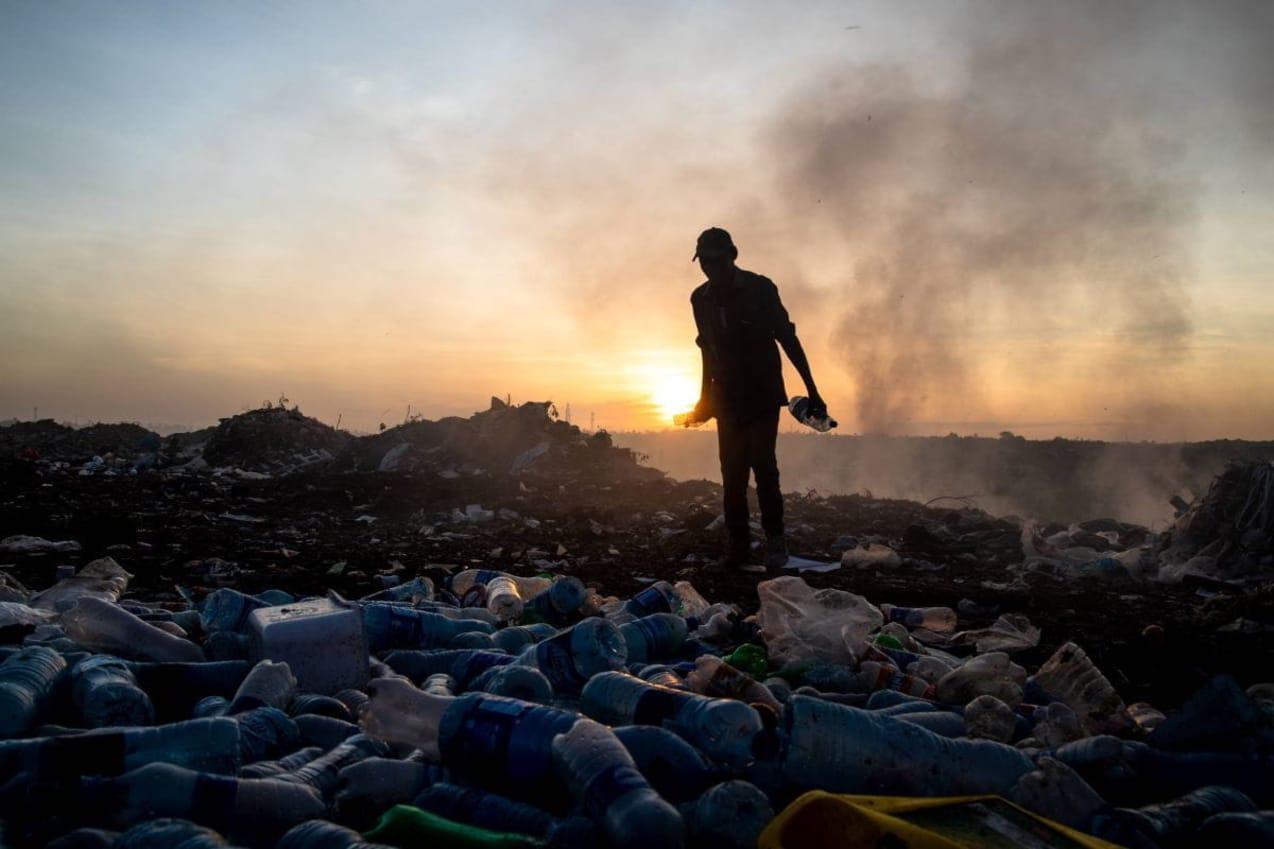A waste picker stands on a pile of waste at sunset. There are plastic bottles in the foreground and smoke rising in the background. 