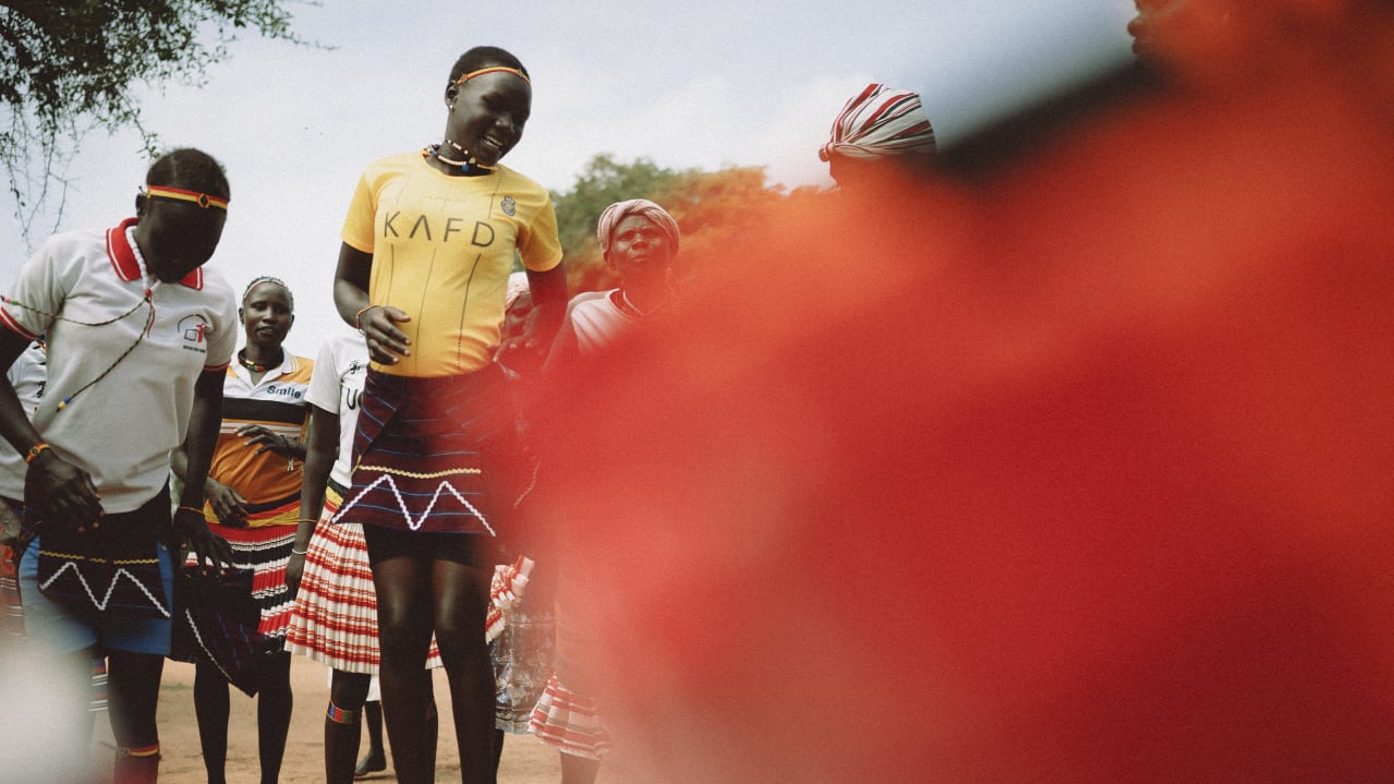 Group of people in traditional and modern attire with beaded skirts and headbands, prominent red blur.