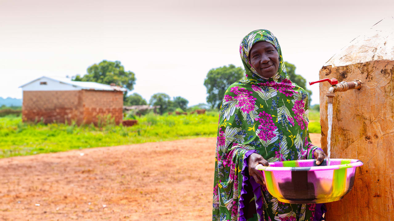 Smiling woman wearing a green and pink floral patterned outfit, holding a multicoloured basin under a flowing outdoor water tap. A small house and trees are visible in the rural background.