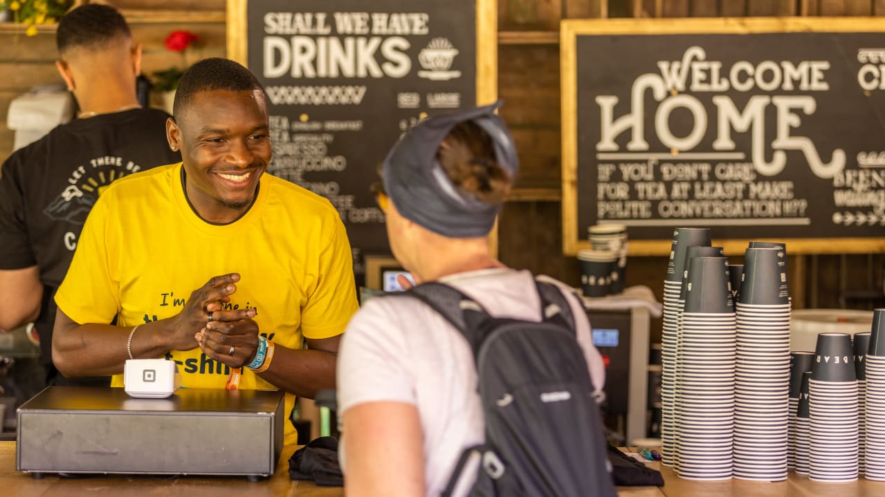 A man dressed in a Tearfund t-shirt smiles at a woman from behind a counter with empty paper cups stacked up.