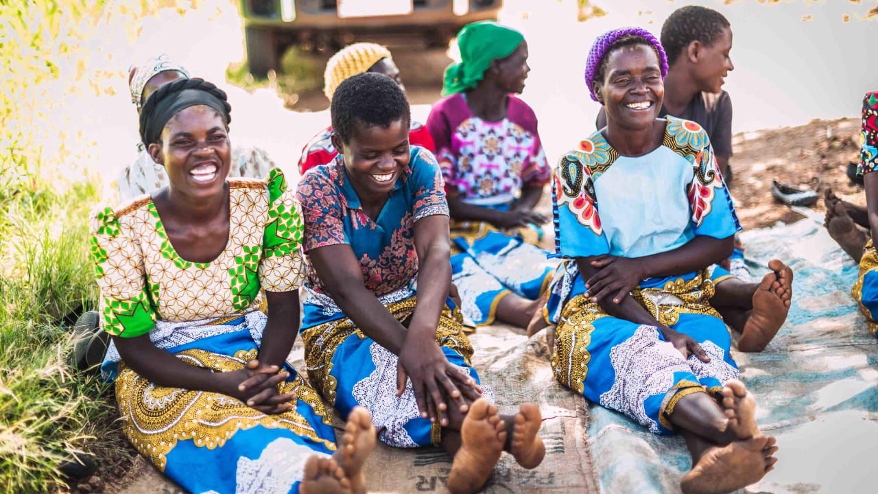 A group of smiling people are sitting on a cloth on the ground outdoors, with a vehicle partially visible in the background. The people in the foreground are barefoot and wearing brightly coloured, patterned clothing.