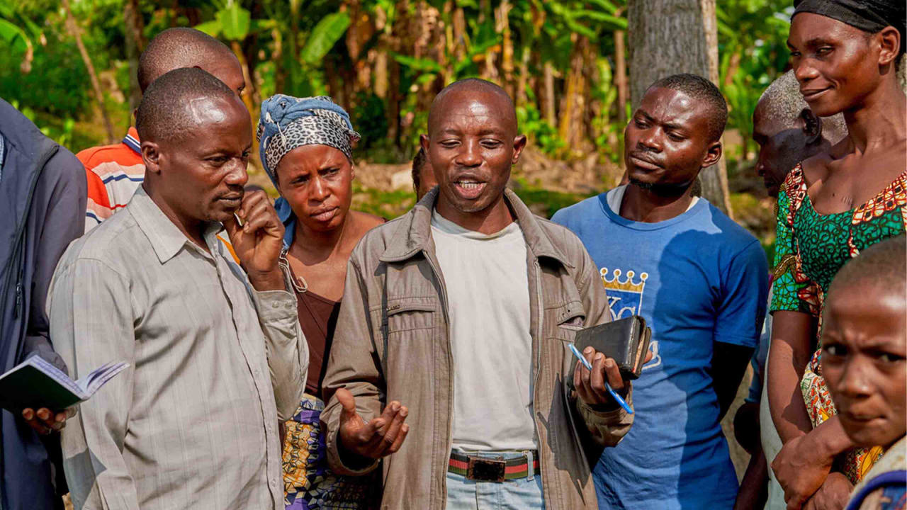 A group of adults, including men and women of varying ages, gather outdoors in a lush, green setting, engaged in a discussion; one man in a light-colored shirt speaks while holding a phone, and others listen attentively.