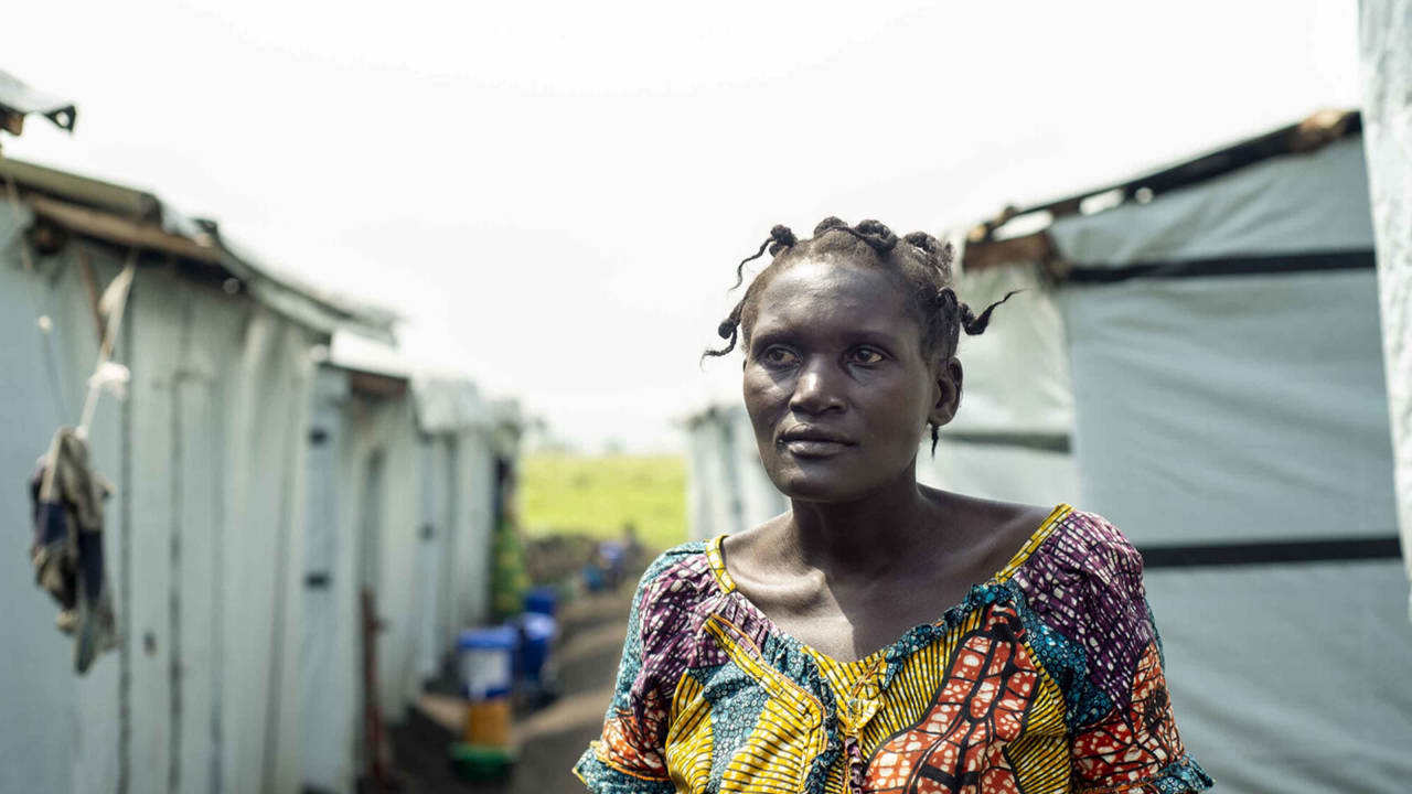 A 35-year-old woman wearing brightly patterned clothes and looking off-camera stands in a path between rows of identical white plastic tents in a refugee camp