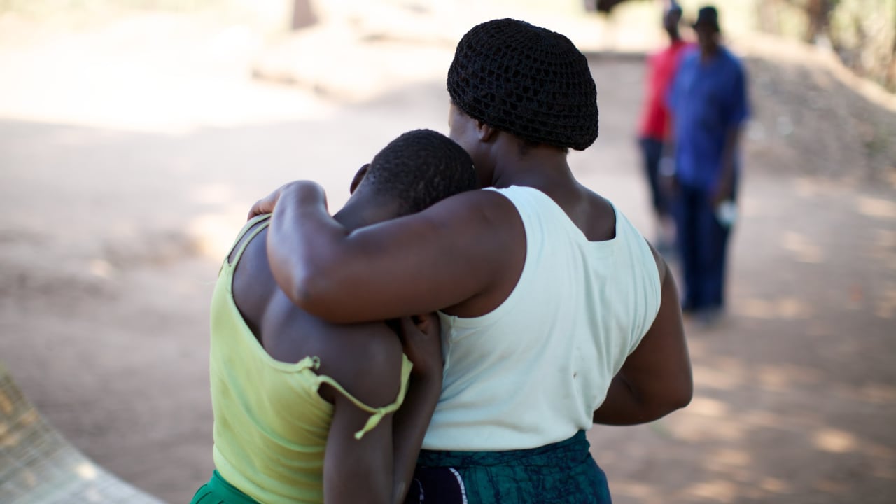 Deux femmes, l’une vêtue d’un haut blanc et l’autre jaune, se tiennent debout l’une serrée contre l’autre.