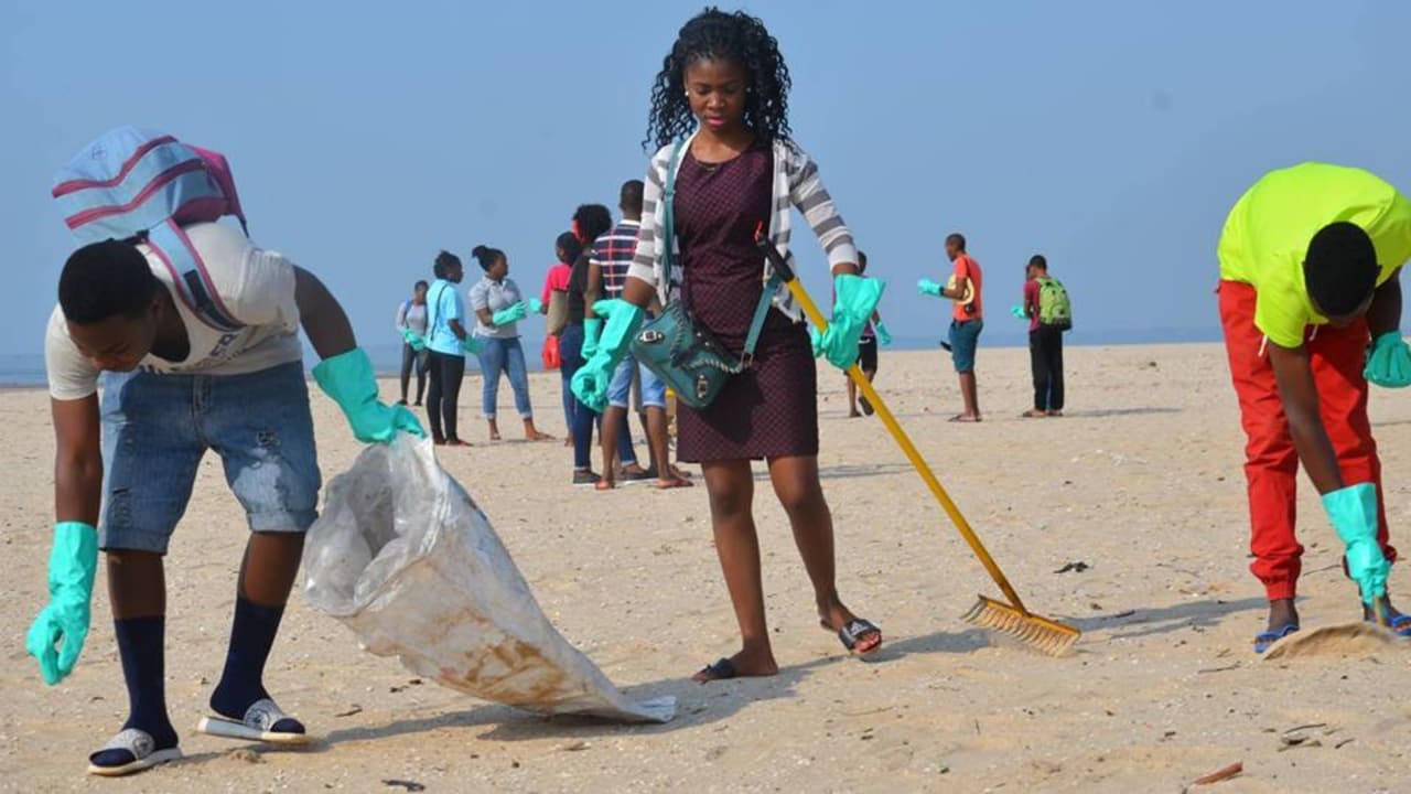 Um grupo de pessoas na praia de Maputo limpando-a com um ancinho e colocando os resíduos em um saco de lixo