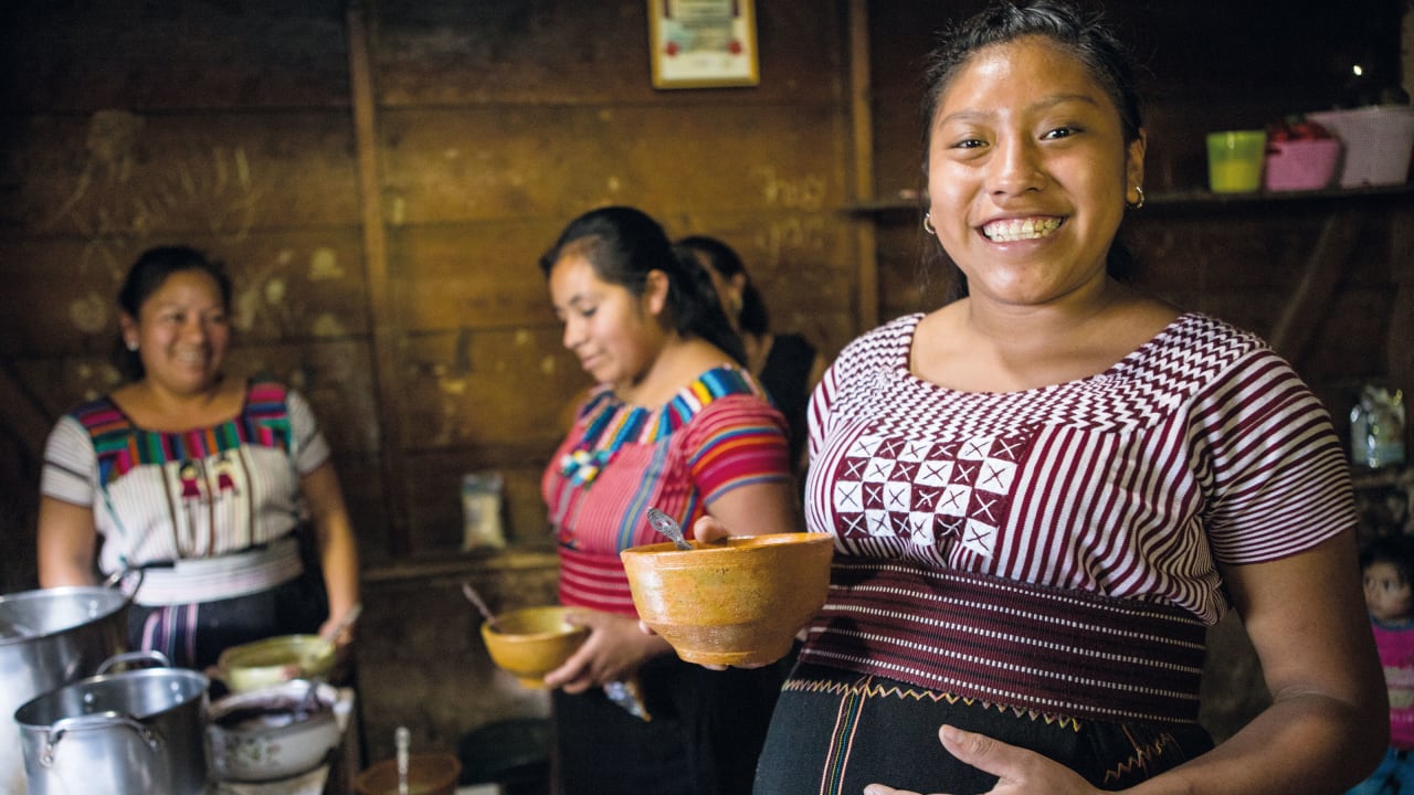 Três mulheres da Guatemala sorrindo, uma delas grávida, segurando tigelas de comida em uma cozinha com paredes de madeira.