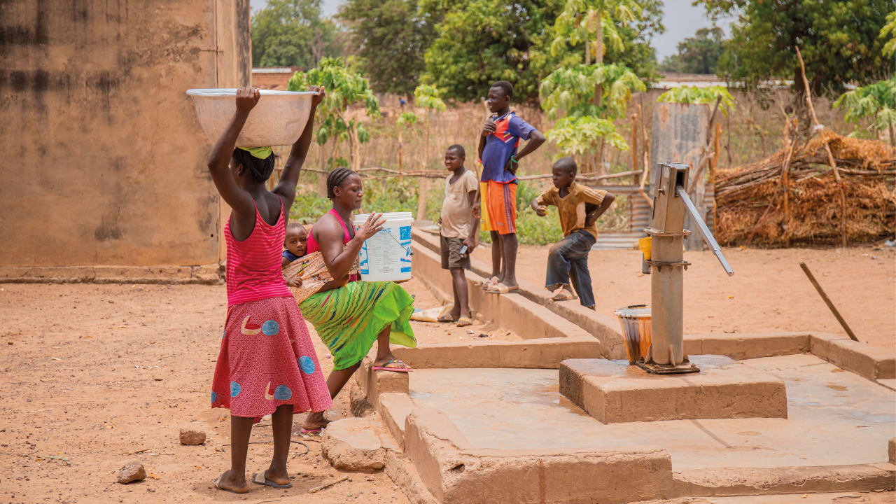 Un grupo de personas jóvenes en torno a una bomba comunitaria de agua y una chica que luce camiseta y falda rosadas y que lleva agua sobre su cabeza en un enorme recipiente