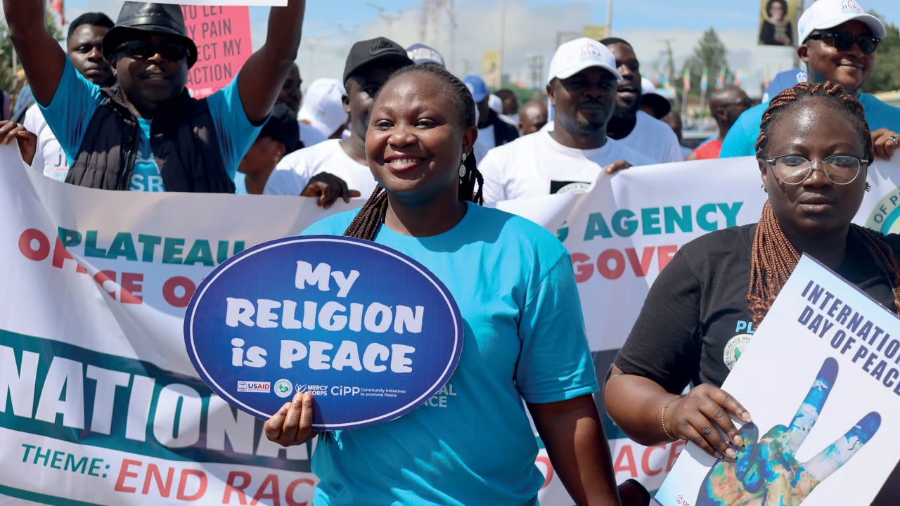 Mulheres e homens nigerianos caminhando em uma rua urbana, segurando cartazes e faixas com mensagens de paz