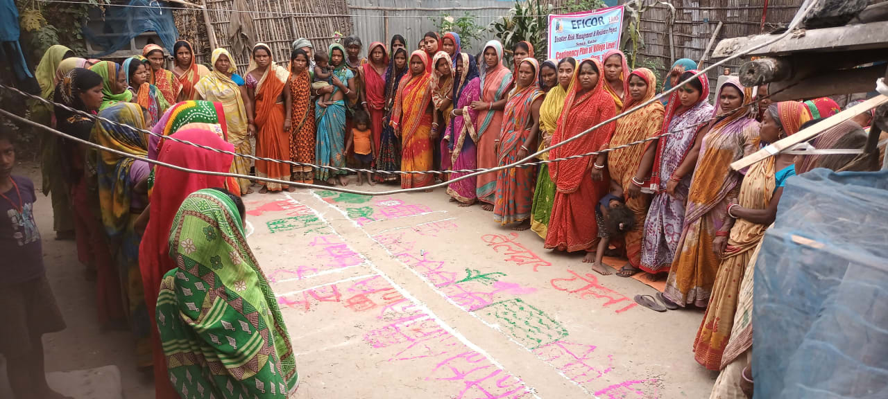 A large group of Indian women dressed in colourful saris stand around the edges of a small courtyard. On the clay floor they have drawn a map of their community using coloured chalk.
