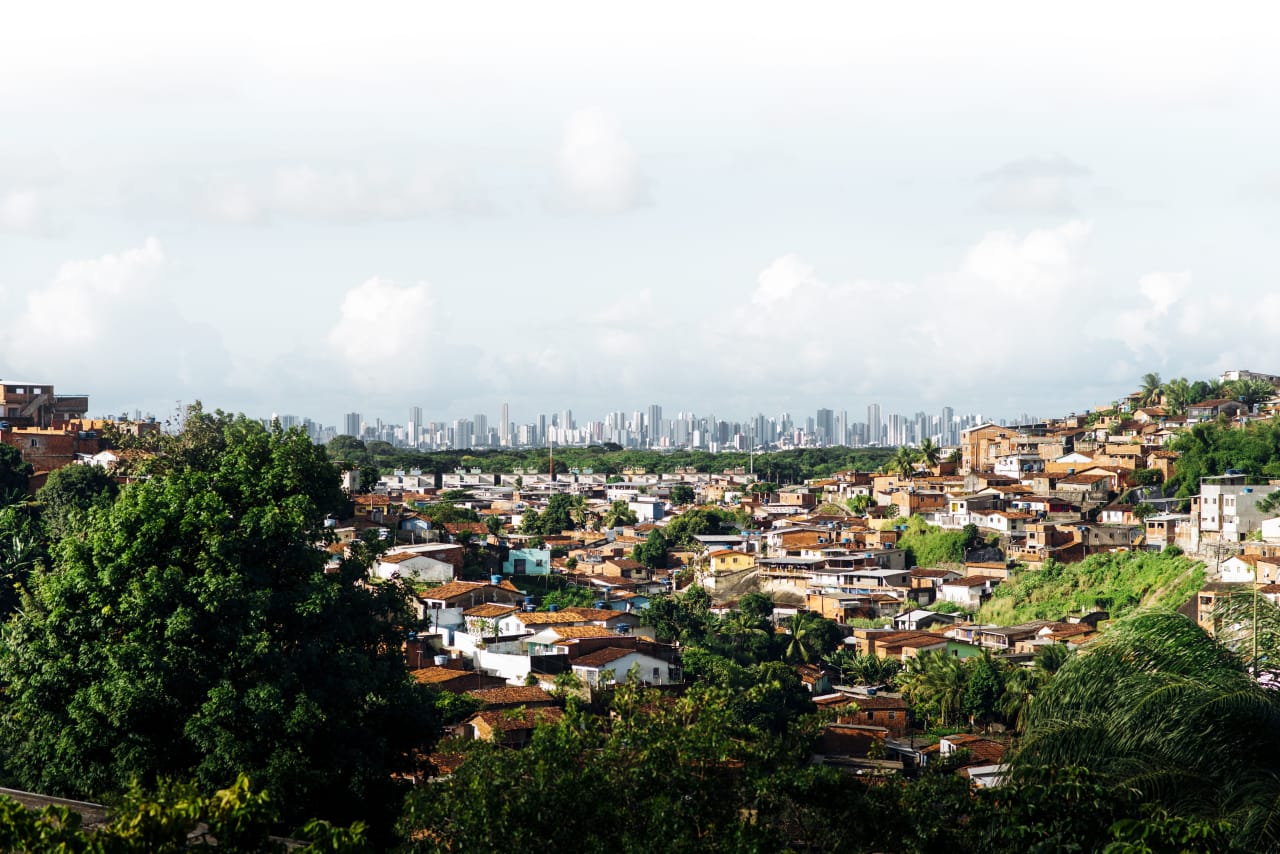 The informal settlement of Sapo Nú stands in the foreground, with the busy city skyline of Recife in the near distance, beyond an area of woodlands.