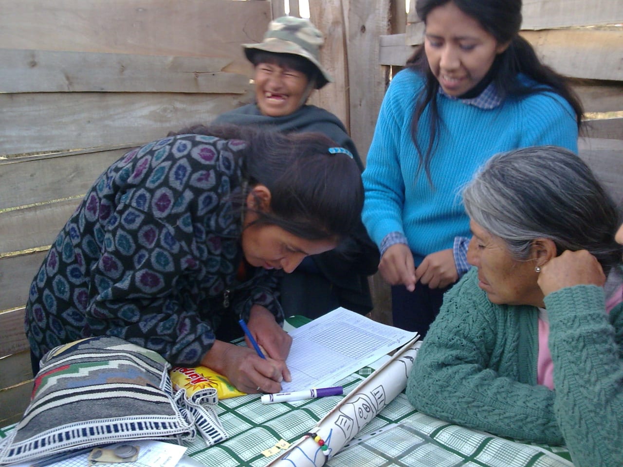 Um grupo de quatro mulheres bolivianas com roupas de inverno reunido em torno de uma mesa, uma delas inclinada e escrevendo em uma folha de registro.