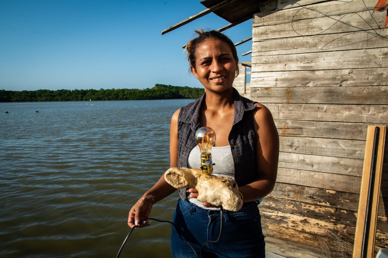 En Colombie, une femme vénézuélienne souriante se tient devant un cabanon en bois qui surplombe la mer, avec un morceau de bois flotté transformé en lampe dans les mains.