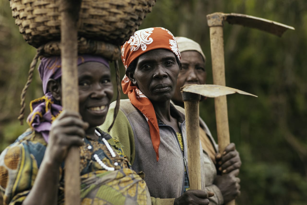 Trois agricultrices rwandaises, l’une avec un panier posé sur sa tête, se tiennent côte à côte avec des binettes à la main, le visage tourné vers l’objectif.
