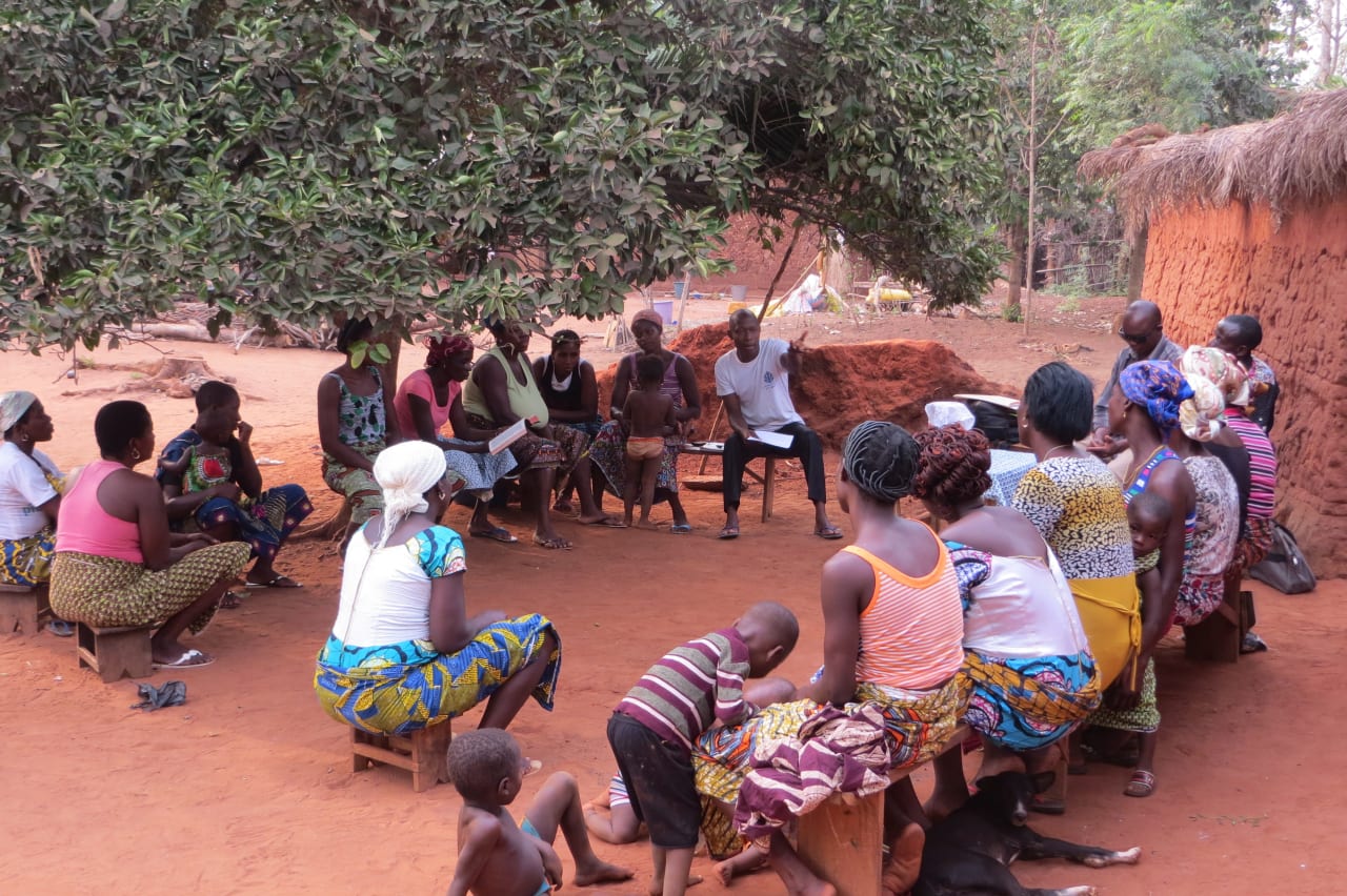 Un numeroso grupo de mujeres, niños y hombres togoleses sentados en bancos de madera bajo un árbol, con casas  hechas de tierra roja al fondo y techos de paja.