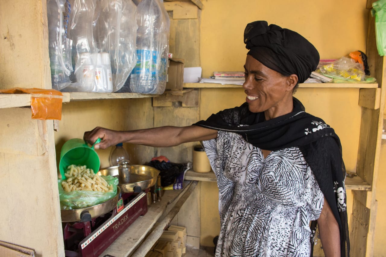 Dans sa petite boutique, une femme éthiopienne souriante vêtue de noir et blanc verse des pâtes dans un sac en plastique vert.