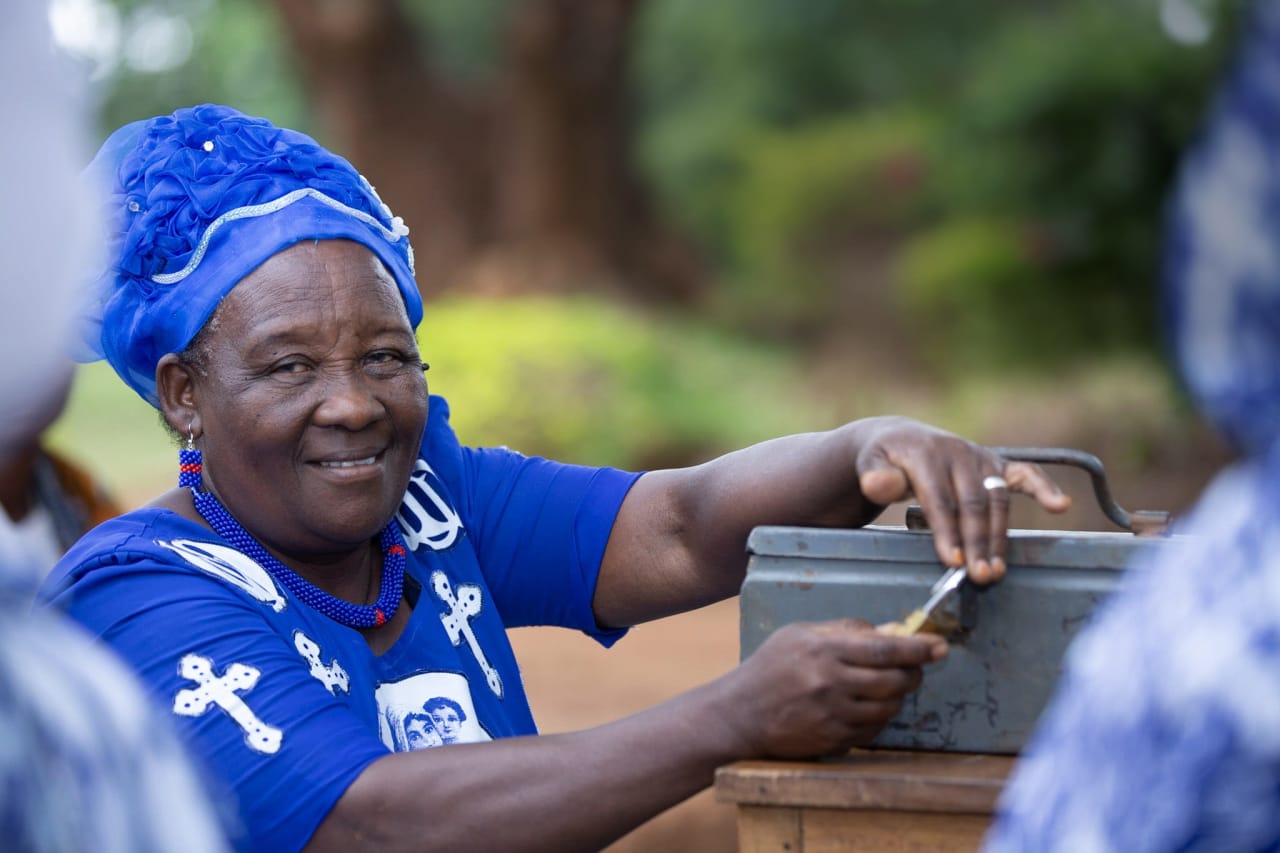A smiling Tanzanian woman wearing a blue and white hat and dress opens the padlock on a grey metal cash box.