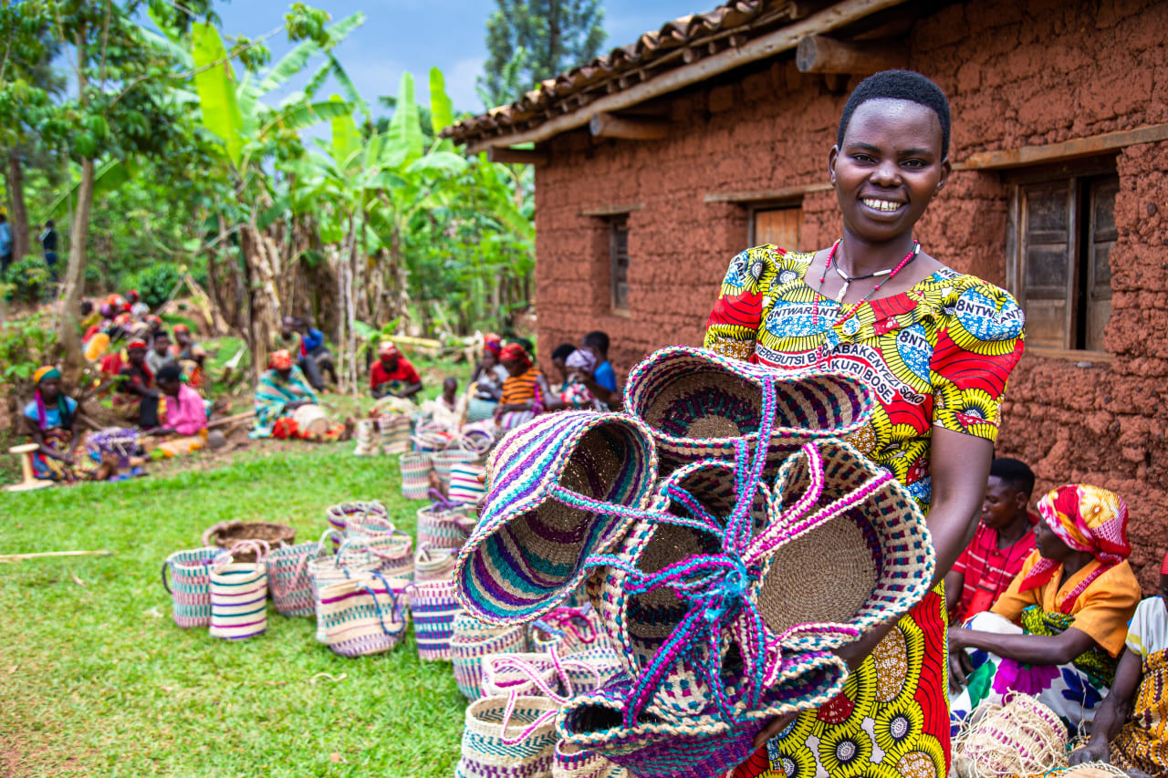 Una mujer burundesa sonriente con un vestido colorido sostiene varias cestas hechas a mano con otros artesanos al fondo.