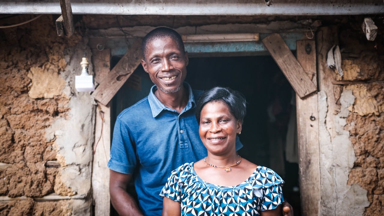 En Côte d’Ivoire, un couple se tient devant l‘entrée de leur maison en souriant à l’objectif.