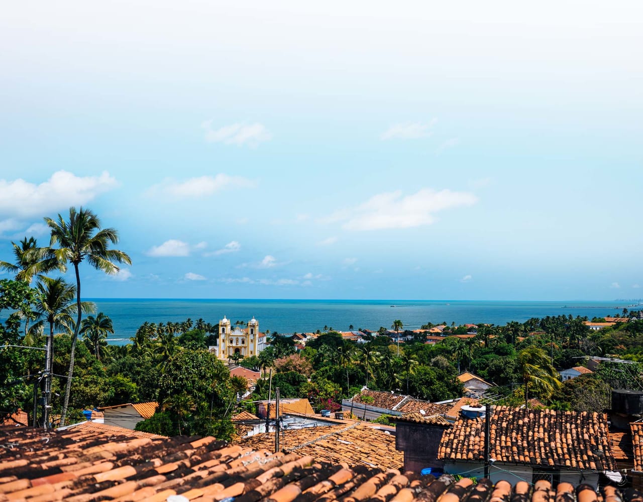 A view over the rooftops of a city in Brazil with a large church in the middle distance and the sea and blue sky behind.