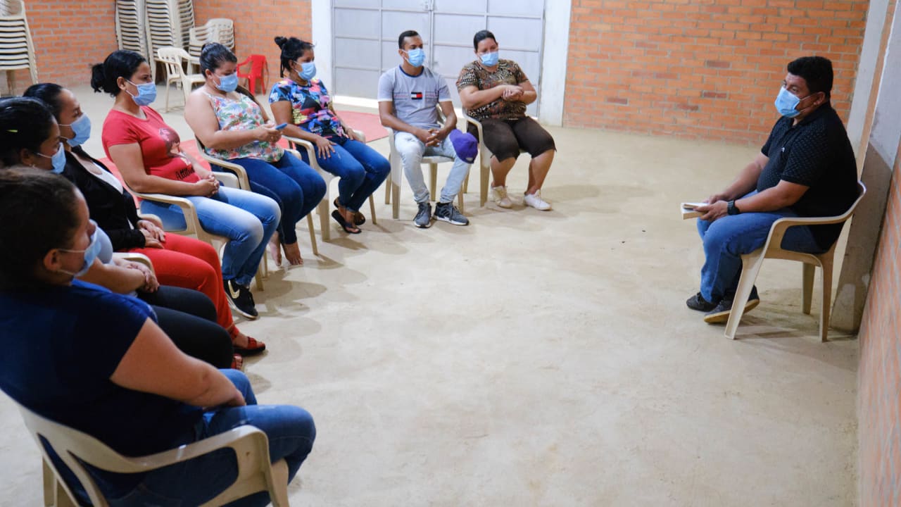 Un groupe d'hommes et de femmes est assis en demi-cercle sur des chaises en plastique alors qu'un homme portant un polo noir les mène lors d'une séance de dévotion.