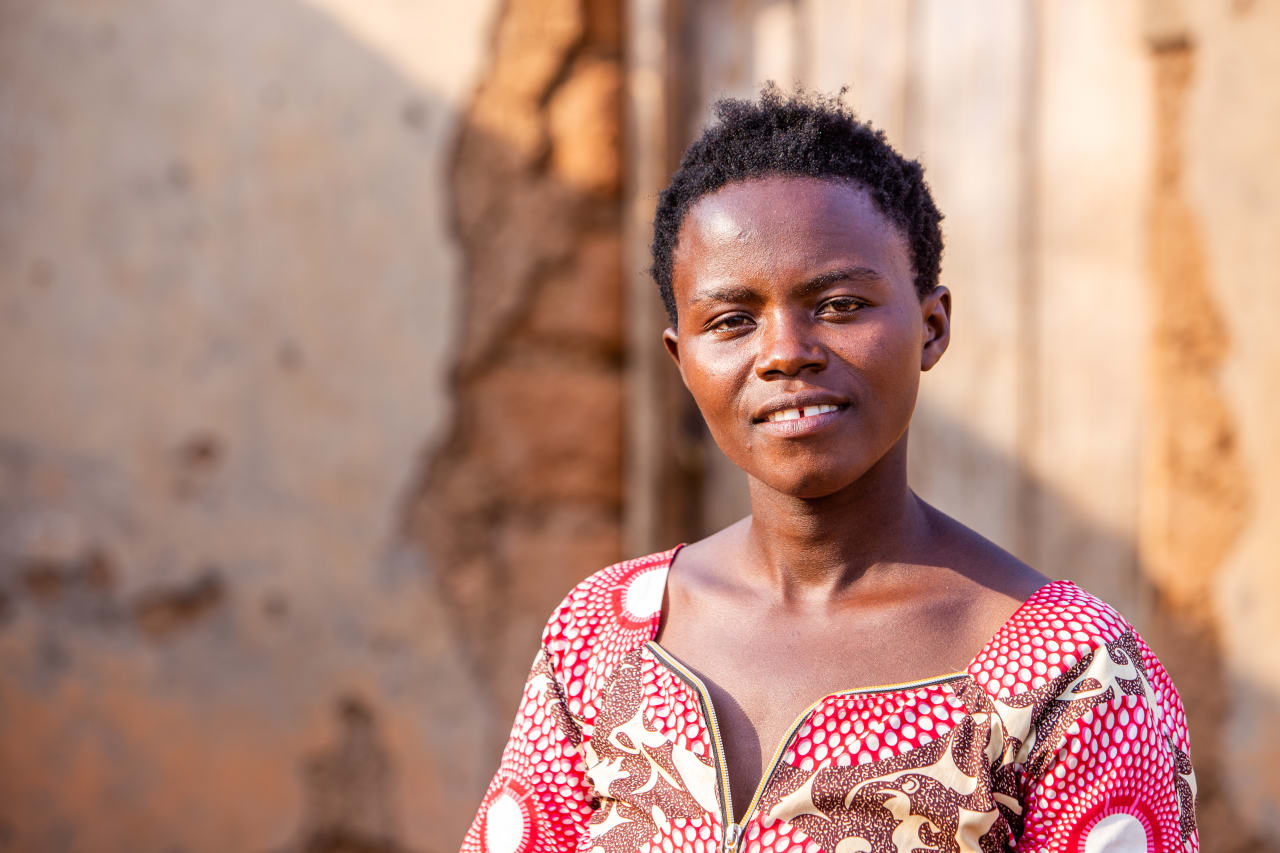  A young woman with short black hair smiles while wearing a colorful patterned dress, standing in front of a weathered wall.