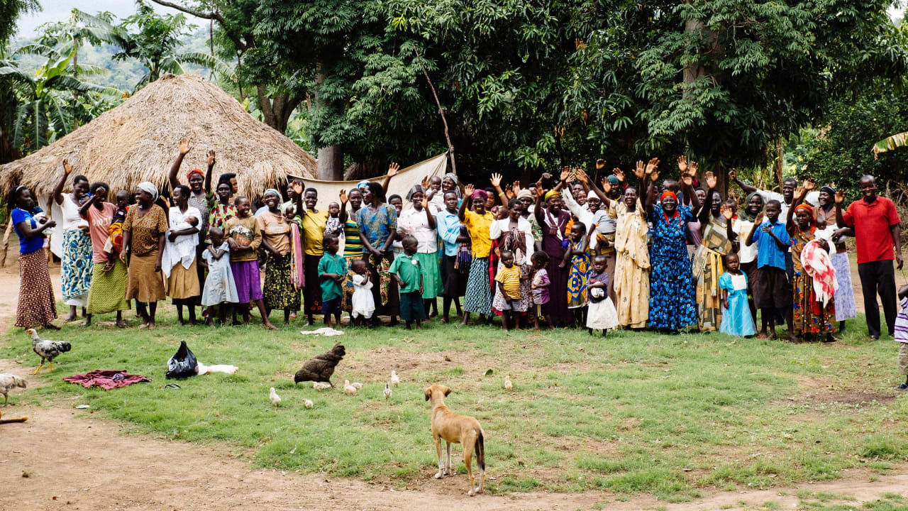 Um grupo da membros de uma comunidade de Uganda – incluindo homens, mulheres, crianças e animais – acenando com as mãos no ar e sorrindo