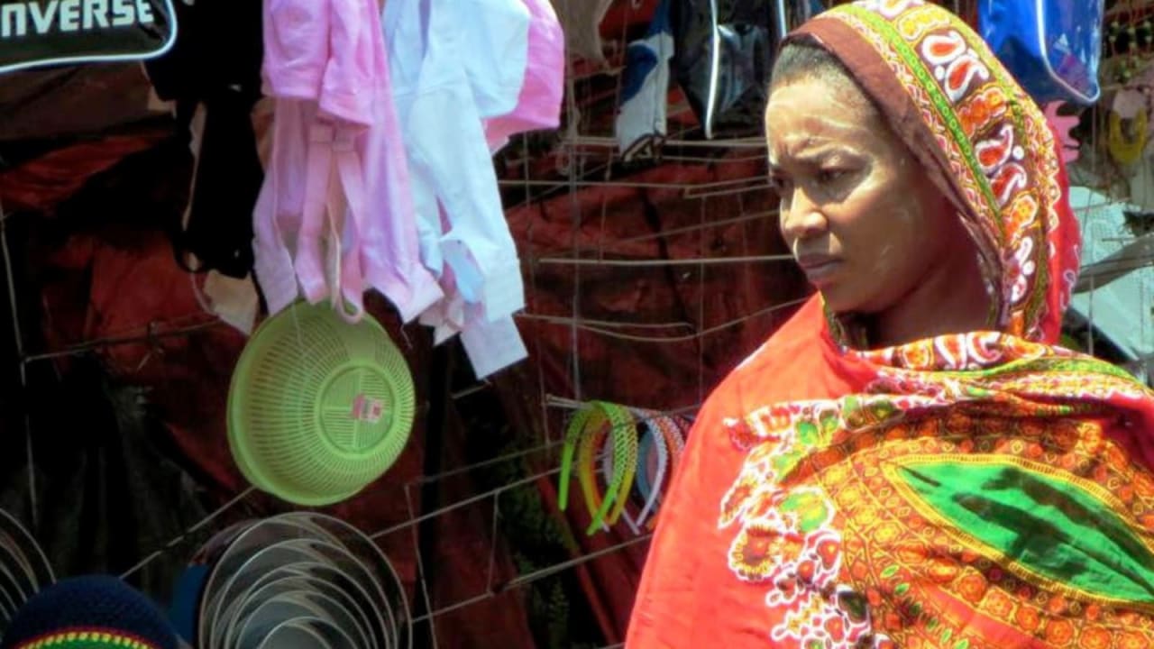 A woman wears a brightly coloured head-covering and stands next to a plastic bowl hanging on a metal fixture