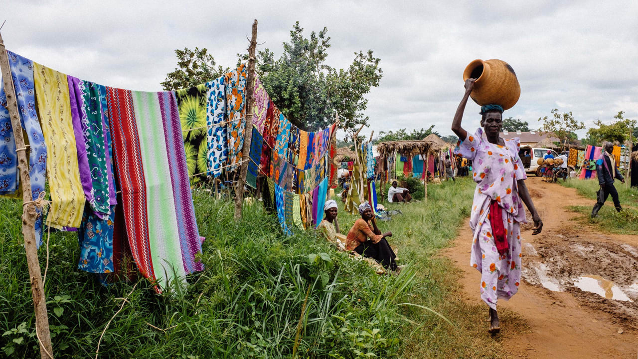 A community in Uganda where brightly coloured fabric is sold at the side of the road and a woman walks past carrying an earthenware pot.