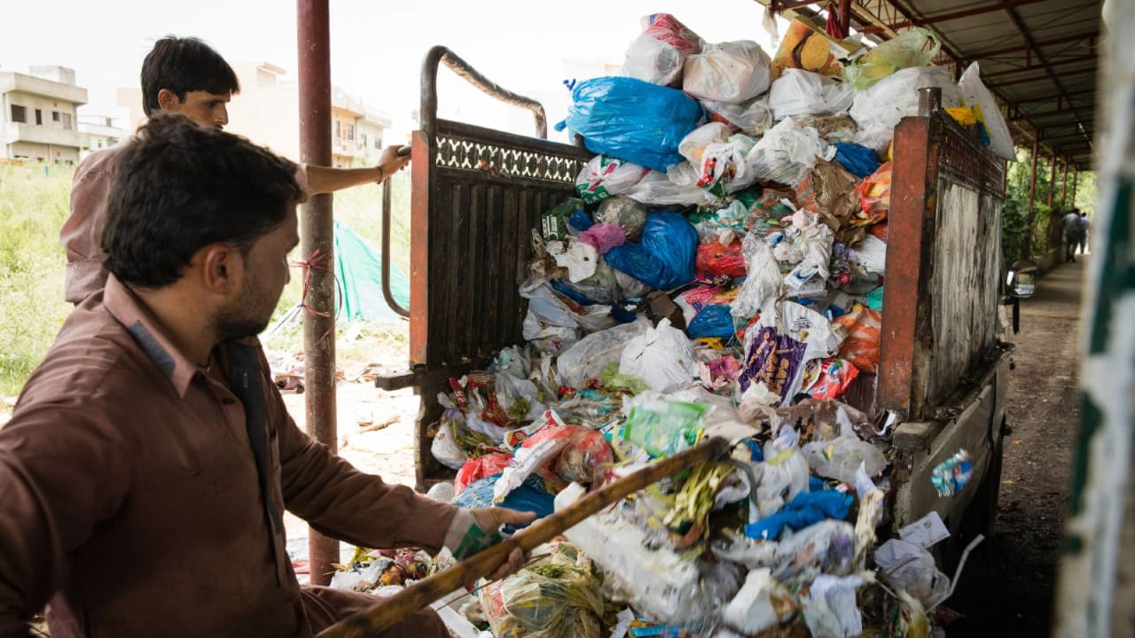 Dois homens em um Centro de Reciclagem de Recursos, em Islamabad, retirando resíduos da traseira de um caminhão com uma ferramenta de madeira