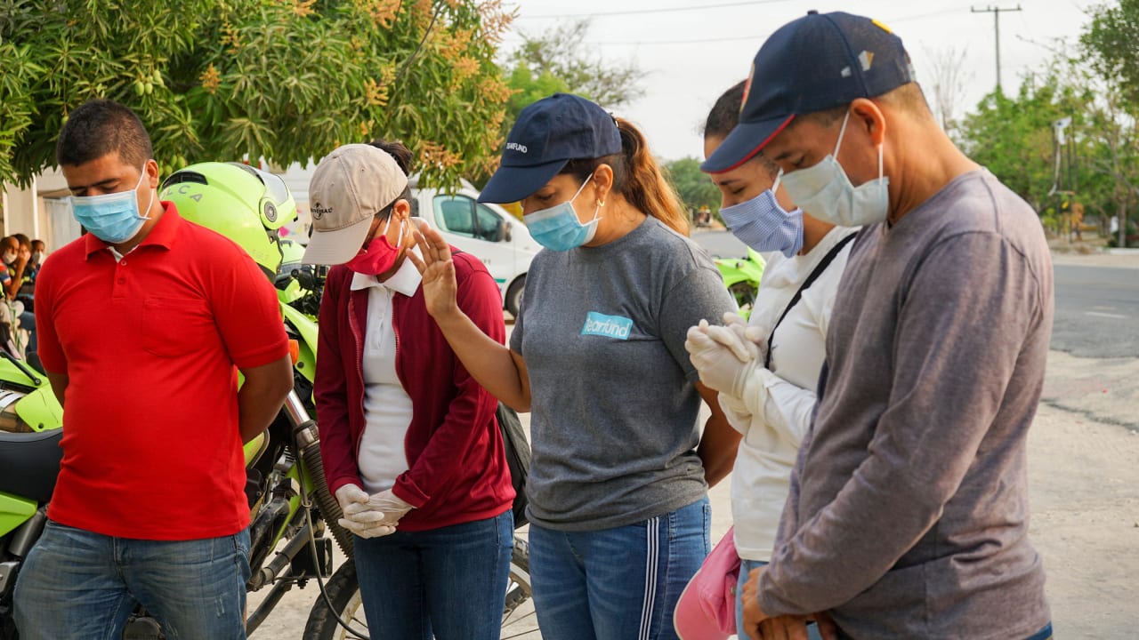 A group of five men and women wearing masks stand on the side of the road praying in preparation for delivering food packages to needy families in Columbia.