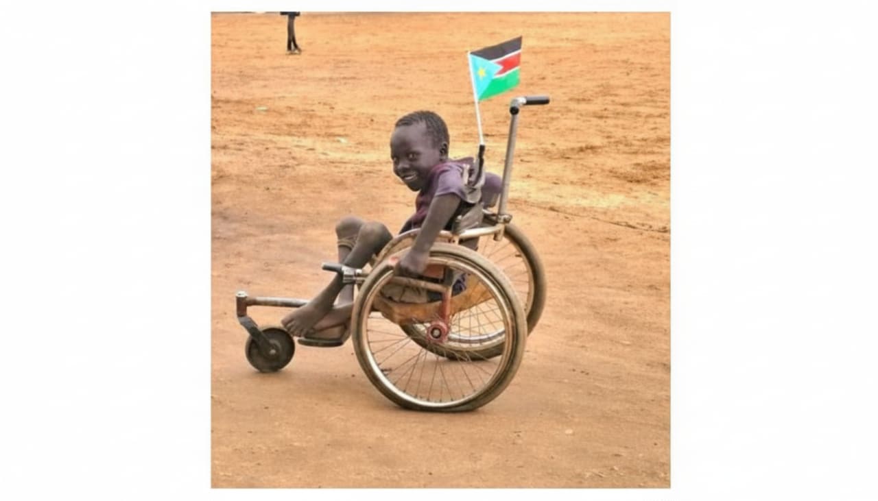 A smiling child sits in a wheelchair, which has a flag attached to the back, on a dirt path with a blurred background.