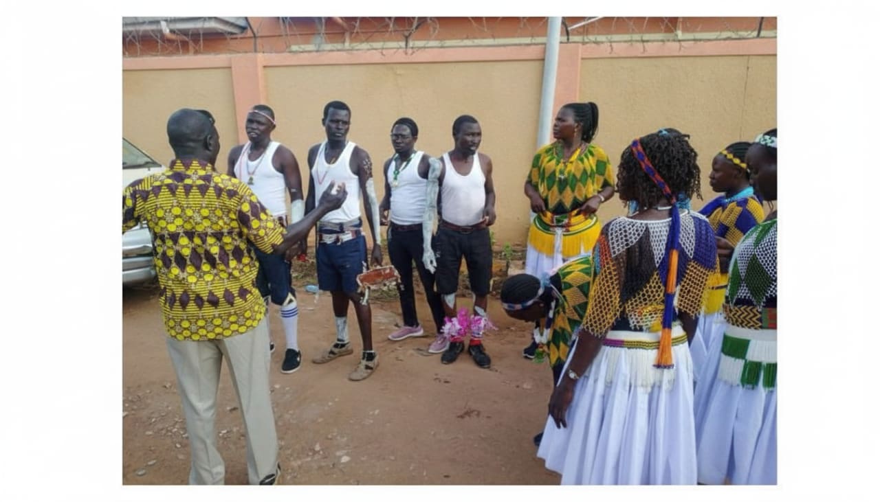 A group of people, some wearing traditional beaded and patterned clothing, are gathered outdoors on a dirt area near a fence, with one person in a yellow patterned shirt facing the group while gesturing.
