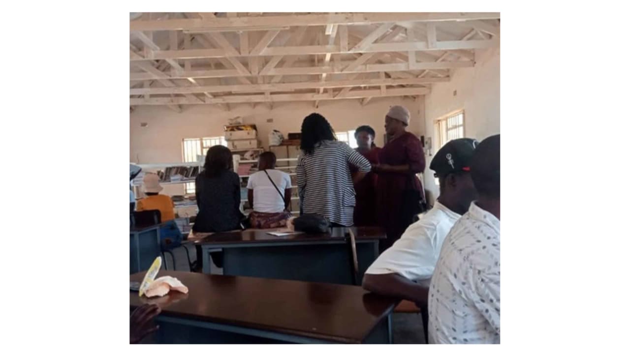 Group of people gathered inside a room with desks and shelves of books, possibly a library.