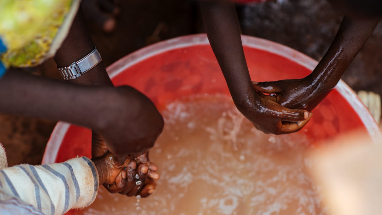 Mãos de duas pessoas, uma delas lavando as mãos de uma criança com sabão sobre um balde de água como parte de uma demonstração de boa higiene em Burundi