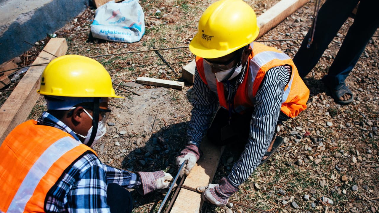 Dois operários da construção civil, usando chapéus amarelos, serrando um pedaço de madeira pelo meio para construir uma casa resistente a terremotos no Nepal
