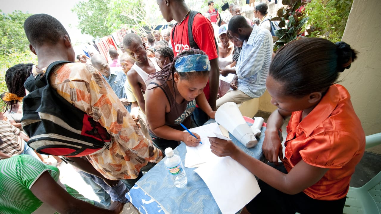 Une femme est assise à un bureau, dans le cadre d’un programme de distribution d’argent en Haïti, où une autre femme signe un papier entourée par la foule.