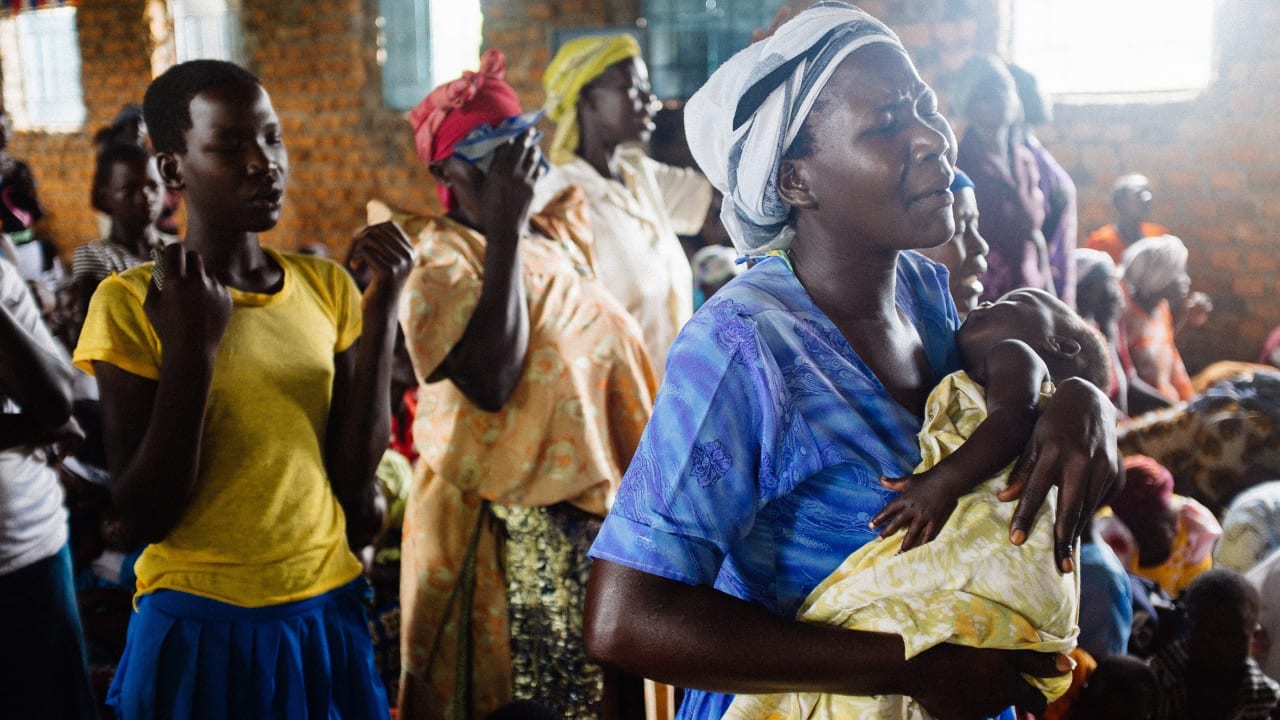 Une femme, vêtue d’une chemise à col bleu, ferme les yeux et tient son bébé pendant qu’elle prie à l’église avec d’autres femmes.