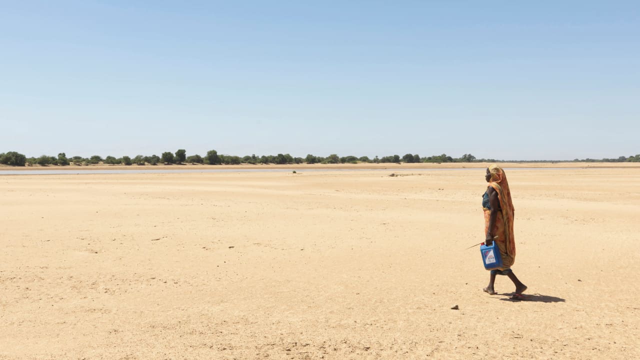 Au Tchad, une femme, portant une jupe et une coiffe orange, marche à la recherche d’eau dans un paysage poussiéreux et sec près du lit d’une rivière asséchée.