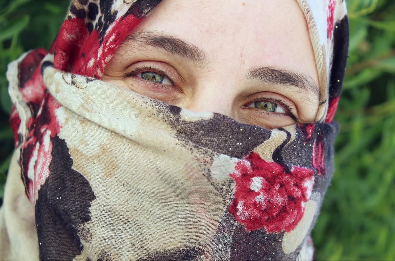 A close-up of a person with light-colored eyes, smiling while wearing a floral-patterned scarf that covers their head and the lower half of their face, set against a blurred green leafy background.